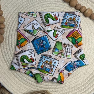 May include: High-waisted baby shorts with a repeating pattern of children's book covers. The books feature illustrations and titles like "The Very Hungry Caterpillar" and "Chicka Chicka Boom Boom." The shorts are made of a soft, ribbed fabric.