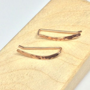 May include: A pair of rose gold ear cuffs with a hammered finish. The cuffs are curved and have a simple, minimalist design.