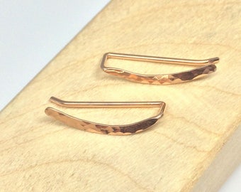 May include: A pair of rose gold ear cuffs with a hammered finish. The cuffs are curved and have a simple, minimalist design.