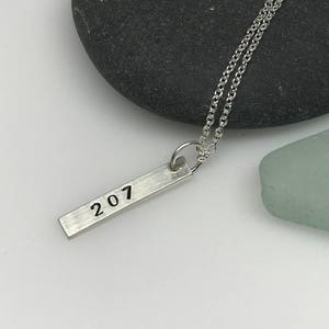 May include: A silver necklace with a silver bar pendant that says "207" on it. The necklace is on a gray rock and a light blue sea glass.