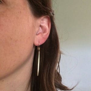 May include: A close-up of a person's ear wearing a pair of gold-colored, long, slender drop earrings. The earrings are straight and have a simple, minimalist design. The background is a soft, neutral color.