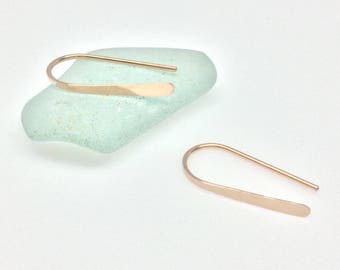 May include: A pair of rose gold minimalist earrings with a simple curved shape. The earrings are made of a thin, flat piece of metal that is bent into a gentle arc.