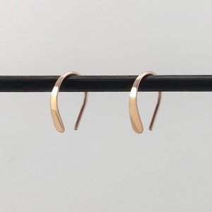 May include: A pair of small, simple, gold hoop earrings. The hoops are made of thin, flat wire and are slightly curved. They are shown hanging from a black rod.