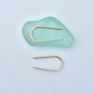 May include: A pair of gold-toned minimalist earrings with a simple curved shape. The earrings are displayed on a piece of light blue sea glass.