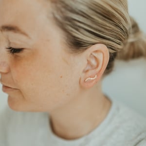 Könnte beinhalten: Nahaufnahme eines Ohrs mit einem kleinen, goldenen, gebogenen Ohrring. Der Ohrring ist ein schlichtes, minimalistisches Design.