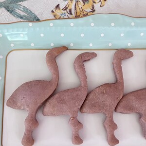May include: Four pink flamingo-shaped cookies on a white plate with a light blue and white polka dot border. The cookies are arranged in a row.