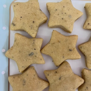 May include: Close-up of star-shaped cookies arranged on a white tray with a light blue and white polka dot border. The cookies are light brown with dark specks, likely herbs or spices, and have a slightly rough texture.