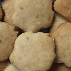 May include: A close-up of a pile of light brown, round, and slightly irregular shaped cookies. The cookies have a slightly rough texture and appear to be homemade.