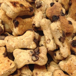 May include: A close-up of a pile of dog biscuits shaped like bones. The biscuits are a light brown color and have chocolate chips in them.