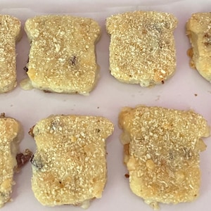 May include: Close-up of several breaded, golden-brown, bite-sized appetizers. The appetizers are shaped like small, rounded rectangles, with a crispy, textured coating. They are arranged on a light pink surface.