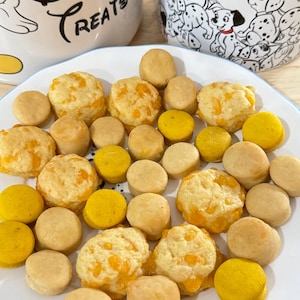 May include: A white plate holds an assortment of baked goods, including golden-yellow and light-brown round treats. Some have a cheesy, orange topping. Two Disney-themed mugs are in the background, one with a Dalmatian pattern and the other with the text "Devon's Treats."