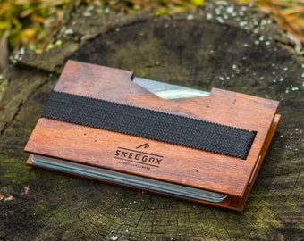 May include: A wooden wallet with a black elastic band and the text "SKEGGÖX" on the front. The wallet is sitting on a tree stump.