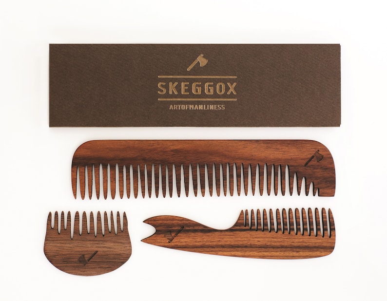 May include: Three wooden combs with a brown wood grain finish. The combs have a small axe logo engraved on them. The combs are arranged on a white background. The text "SKEGGOX ARTOFMANLINESS" is printed on a brown card behind the combs.
