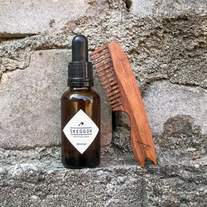 May include: A brown glass bottle with a black dropper top and a white label that reads "SKEGG&Ouml;X ART OF MANLINESS 30ml (1 fl oz)". A wooden comb with a brown handle is next to the bottle.