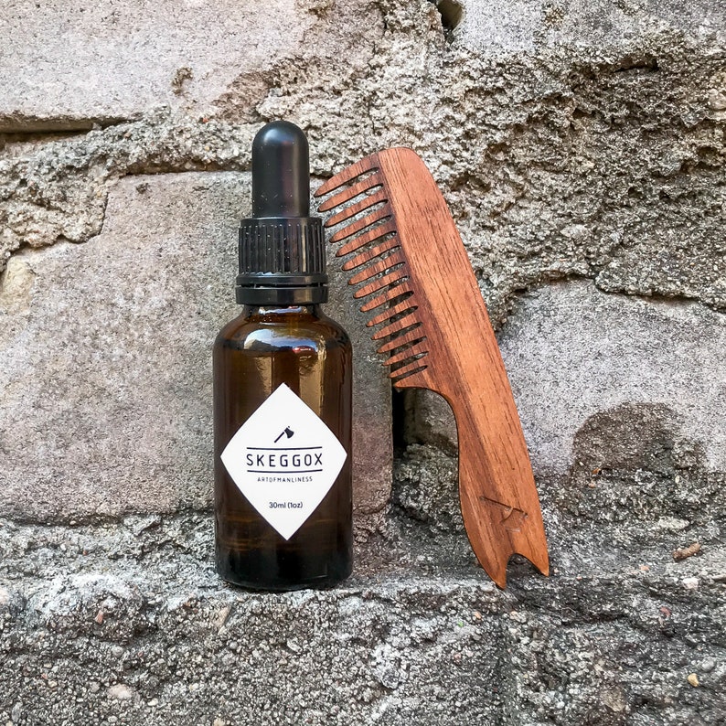 May include: A brown glass bottle with a black dropper top and a white label that reads "SKEGG&Ouml;X ART OF MANLINESS 30ml (1 fl oz)". A wooden comb with a brown handle is next to the bottle.