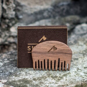 May include: A wooden comb with a rounded top and a small axe design carved into the wood. The comb is sitting on a brown leather box with the word "SK" visible.