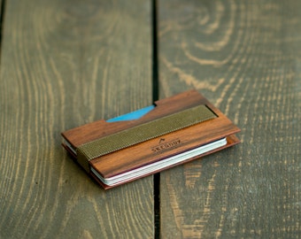 May include: A wooden wallet with a brown leather strap and the text "Skeegox" engraved on the front. The wallet is closed and lying on a wooden surface.