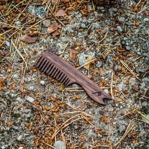 May include: A wooden comb with a dark brown finish and a small bottle opener on the end. The comb is lying on a bed of brown pine needles and small rocks.