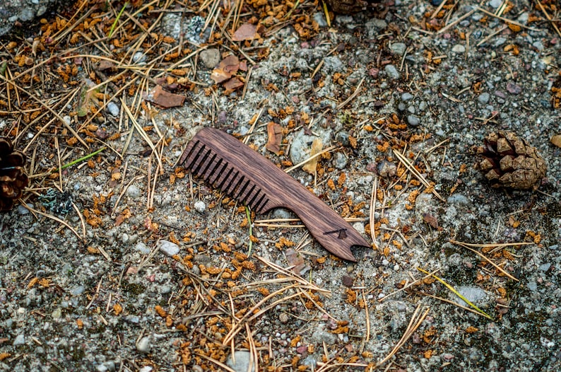 May include: A wooden comb with a dark brown finish and a small bottle opener on the end. The comb is lying on a bed of brown pine needles and small rocks.