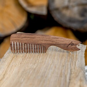 May include: A wooden comb with a brown finish and a small axe design on the handle. The comb is lying on a piece of wood.