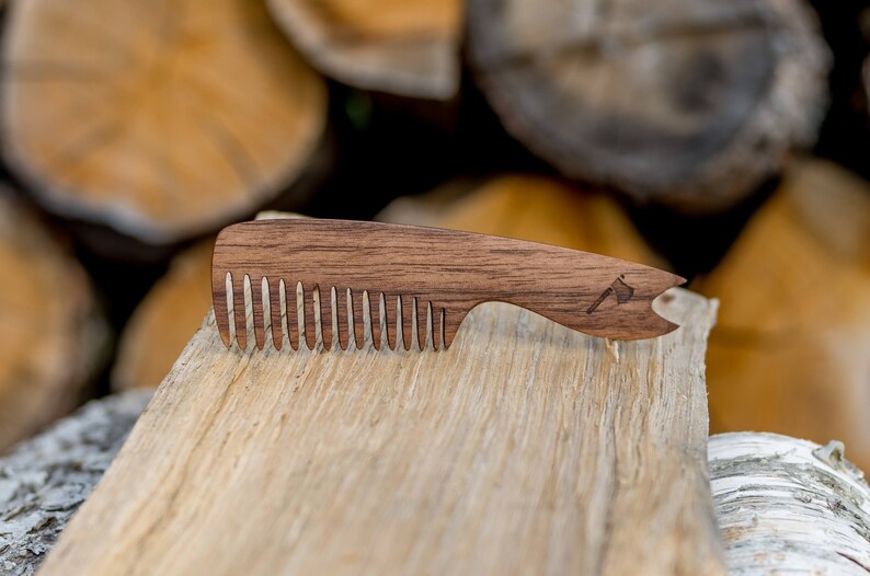 May include: A wooden comb with a brown finish and a small axe design on the handle. The comb is lying on a piece of wood.