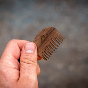 May include: A wooden comb with a rounded shape and a small axe design carved into the handle. The comb has fine teeth and is held in a hand.