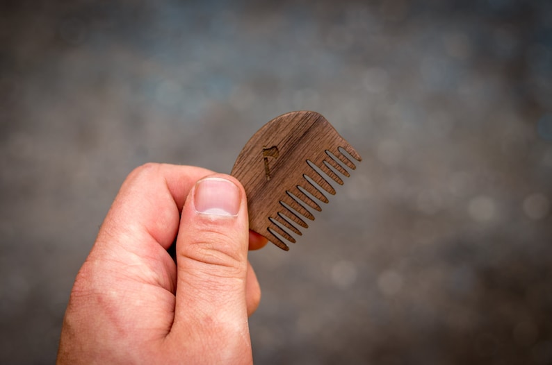 May include: A wooden comb with a rounded shape and a small axe design carved into the handle. The comb has fine teeth and is held in a hand.