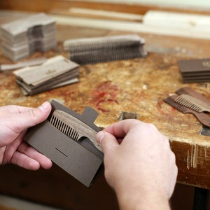 May include: A person holds a brown wooden comb in a brown cardboard sleeve. The comb has a handle with a small notch at the end. The comb is on a wooden table with other combs and packaging.