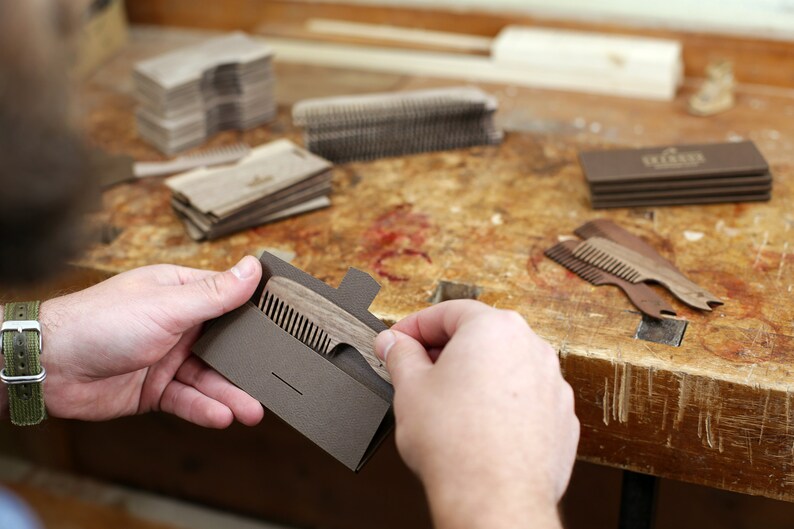 May include: A person holds a brown wooden comb in a brown cardboard sleeve. The comb has a handle with a small notch at the end. The comb is on a wooden table with other combs and packaging.