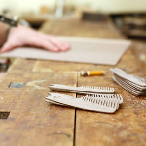May include: A close-up of a wooden workbench with several wooden combs laid out on the surface. The combs are made of light-colored wood and have a natural finish. The workbench is made of dark wood and has a worn surface.