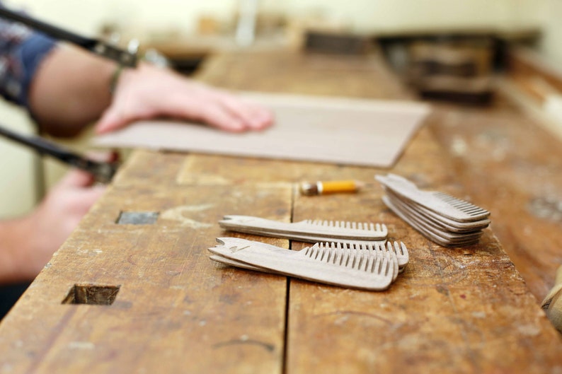 May include: A close-up of a wooden workbench with several wooden combs laid out on the surface. The combs are made of light-colored wood and have a natural finish. The workbench is made of dark wood and has a worn surface.