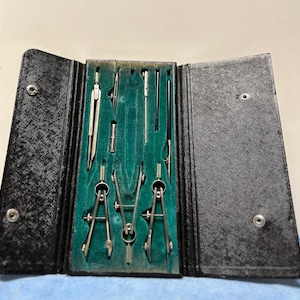 May include: A black leather case with a green felt interior holds a set of drafting tools, including compasses, rulers, and a pencil.