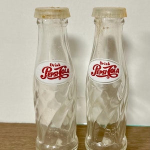 May include: Two vintage Pepsi-Cola glass bottles. The clear bottles have a twist design and are capped. Each bottle has a white and red Pepsi-Cola logo. The bottles are displayed on a wooden surface.