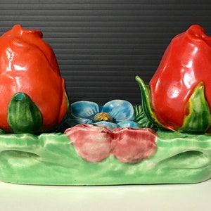 May include: A ceramic salt and pepper shaker set shaped like red roses with green leaves. The shakers are sitting on a green leaf-shaped tray with a blue and pink flower.