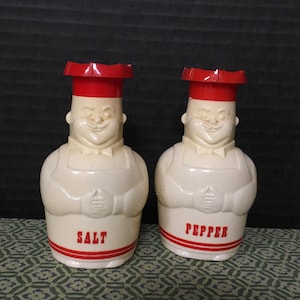 May include: A set of two ceramic salt and pepper shakers shaped like chefs wearing red hats. The shakers are white with red accents and the words "SALT" and "PEPPER" are printed on the front.