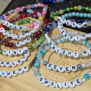 Op de afbeelding: Een verzameling kralenarmbanden in verschillende kleuren, waaronder roze, groen en blauw. Sommige armbanden hebben witte letterkralen met woorden als "LOVER", "FEARLESS" en "WILLOW". De armbanden liggen op een houten oppervlak.