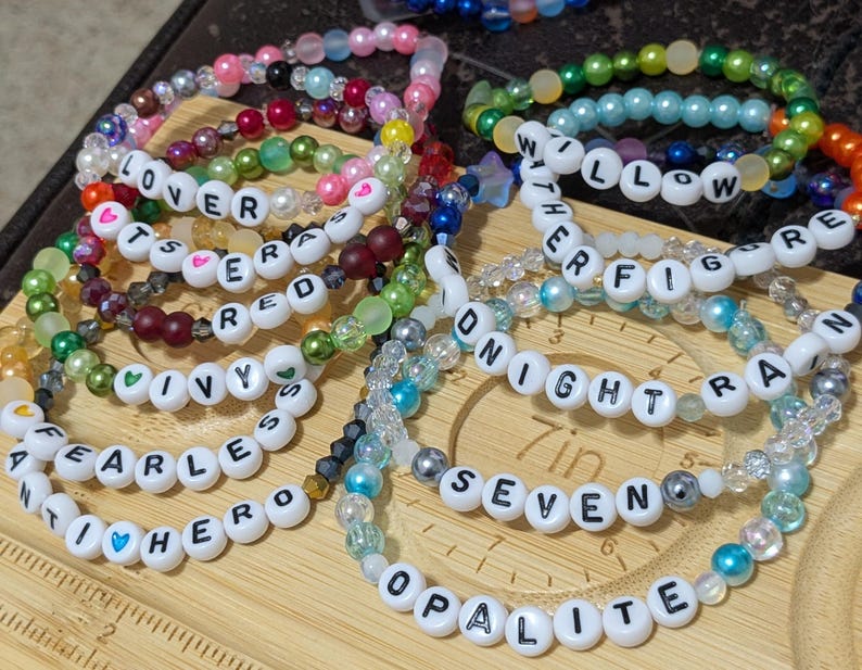Op de afbeelding: Een verzameling kralenarmbanden in verschillende kleuren, waaronder roze, groen en blauw. Sommige armbanden hebben witte letterkralen met woorden als "LOVER", "FEARLESS" en "WILLOW". De armbanden liggen op een houten oppervlak.