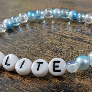Op de afbeelding: Een kralenarmband met het woord "OPALITE" in witte letterkralen. De armband heeft heldere, lichtblauwe en iriserende kralen. De armband ligt op een donkerhouten oppervlak. De kralen zijn ongeveer 0,8 cm in diameter.