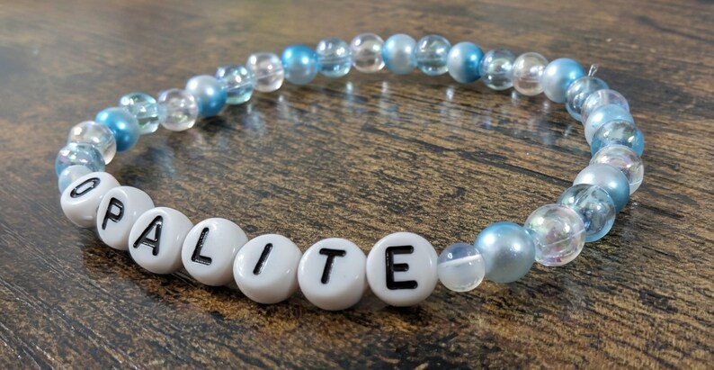 Op de afbeelding: Een kralenarmband met het woord "OPALITE" in witte letterkralen. De armband heeft heldere, lichtblauwe en iriserende kralen. De armband ligt op een donkerhouten oppervlak. De kralen zijn ongeveer 0,8 cm in diameter.