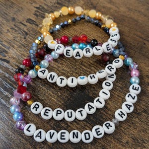 Op de afbeelding: Vier armbanden met kralen met witte letterkralen met de woorden "FEARLESS", "ANTIHERO", "REPUTATION" en "LAVENDER HAZE". De armbanden hebben een verscheidenheid aan kleurrijke kralen in tinten goud, blauw, rood, roze en paars.