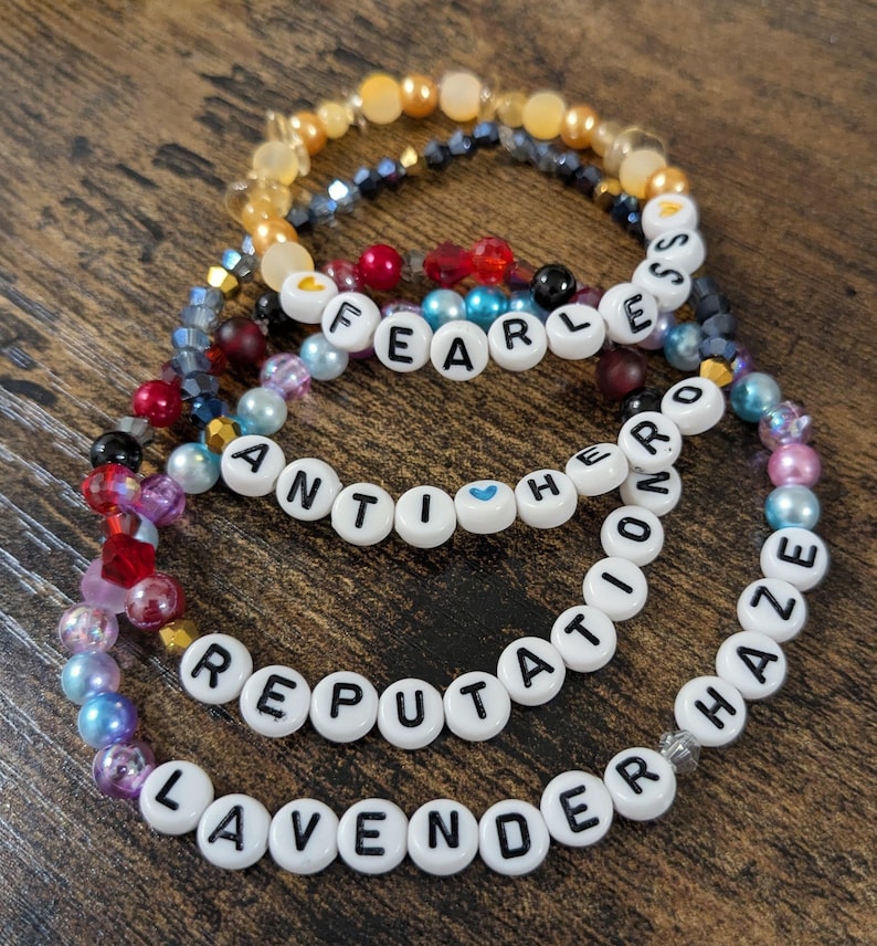 Op de afbeelding: Vier armbanden met kralen met witte letterkralen met de woorden "FEARLESS", "ANTIHERO", "REPUTATION" en "LAVENDER HAZE". De armbanden hebben een verscheidenheid aan kleurrijke kralen in tinten goud, blauw, rood, roze en paars.