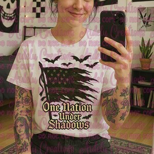 Könnte beinhalten: Weißes T-Shirt mit einem verwitterten amerikanischen Flaggen-Design, Fledermäusen und dem Text "One Nation Under Shadows" in einer gotischen Schrift. Das Shirt hat einen Rundhalsausschnitt und kurze Ärmel.
