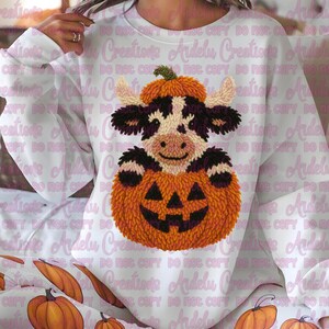 以下が含まれることがあります： ハロウィーンをテーマにしたデザインの白いスウェットシャツ。黒と白の模様の牛が、カボチャの帽子をかぶり、彫刻されたオレンジ色のカボチャを持っています。季節の衣料品です。