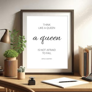 May include: A framed print with the quote "THINK LIKE A QUEEN a queen IS NOT AFRAID TO FAIL" by Oprah Winfrey. The print is in a dark brown frame, set on a wooden desk with plants, a lamp, and books.