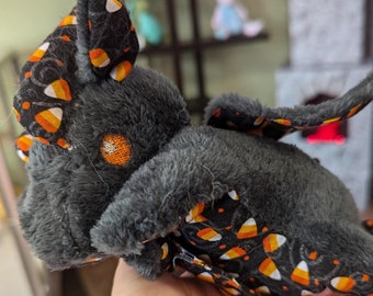 Puede incluir: Un peluche de murciélago con pelaje gris y ojos bordados en naranja. Las orejas y alas del murciélago presentan una tela negra estampada con caramelos de maíz. Un peluche con temática de Halloween.