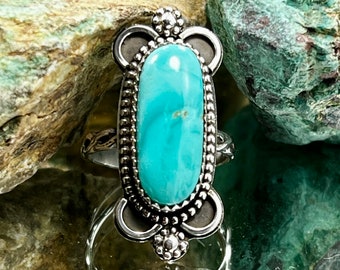 May include: A silver ring with a turquoise stone set in a decorative oval bezel. The ring has a detailed design with small beads and a scalloped edge.