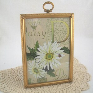 May include: A rectangular gold-toned metal picture frame with an ornate border and a top hanging loop. The frame contains a decorative print featuring white daisy flowers, green leaves, a small ladybug, and the text Daisy D and Da.