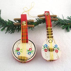 May include: Two wooden musical instrument ornaments. One is round, the other is teardrop-shaped. Both have red tops, cream-colored bodies, and are decorated with floral designs in pink and blue. Gold strings and brown pegs complete the design.