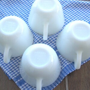May include: Four white, vintage Fire King cups with handles are arranged on a blue and white checkered cloth. The cups are round with a smooth, glossy finish. The handles are a simple, curved design.