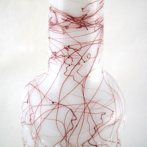 May include: A white glass vase featuring a narrow neck and a rounded body, decorated with a pattern of thin, dark red lines that crisscross across the surface in a chaotic, hand-drawn style, creating a stark contrast against the opaque white glass.